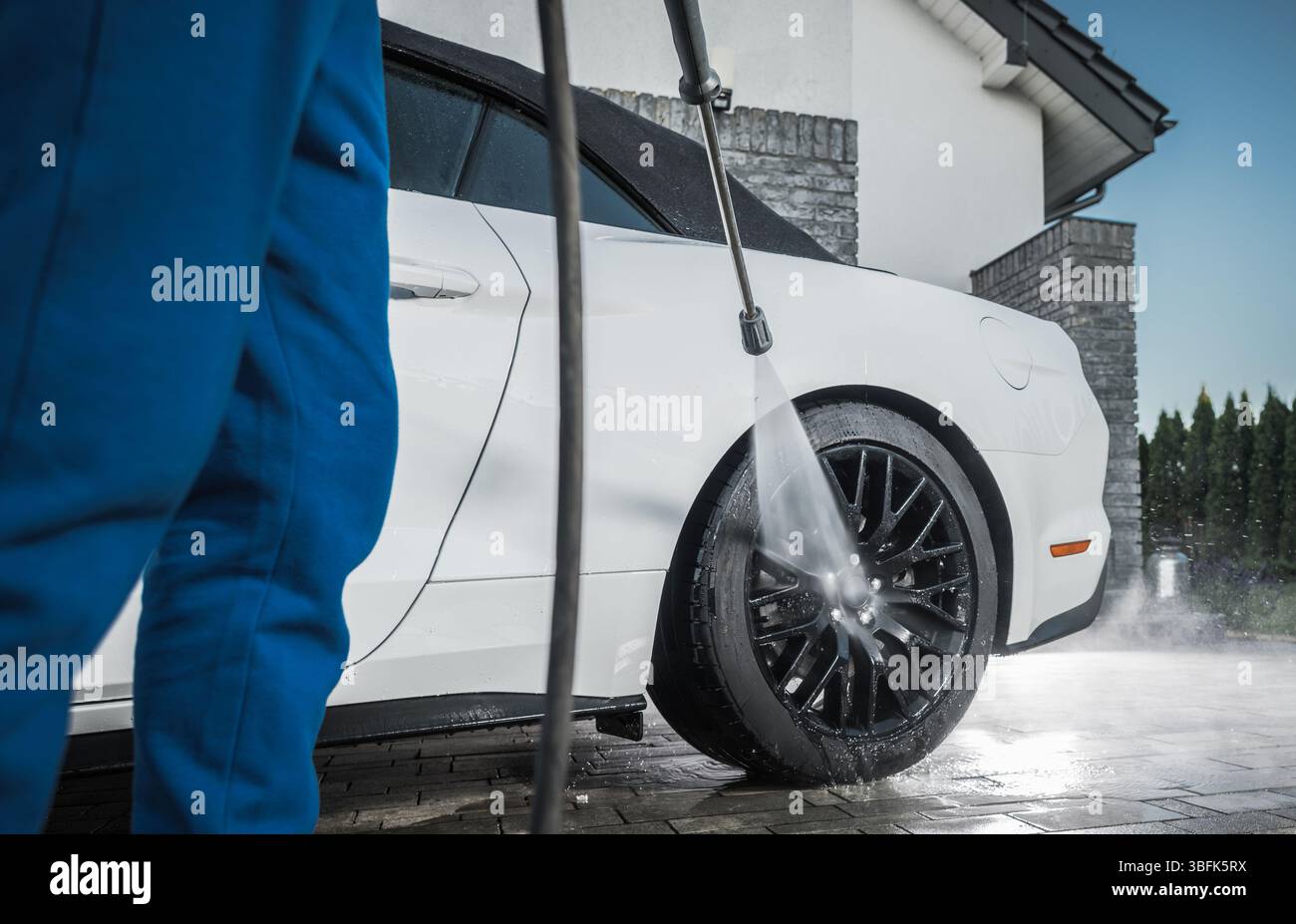 Eine Person sprüht Wasser auf ein weißes Auto in einer Auffahrt, wobei sie sich auf die Räder konzentriert. Die Umgebung bietet ein gut gepflegtes Haus und Grün. Stockfoto