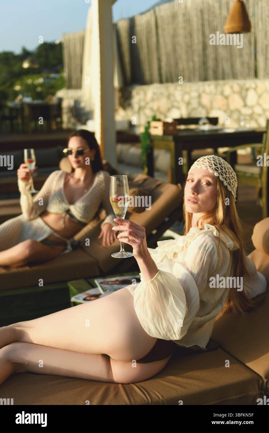 Eine Brünette und Blondine in ihrer Lounge aus den 20er Jahren auf den Liegen am Hotelpool und genießen während der goldenen Sonnenuntergangsstunde Champagner. Stockfoto