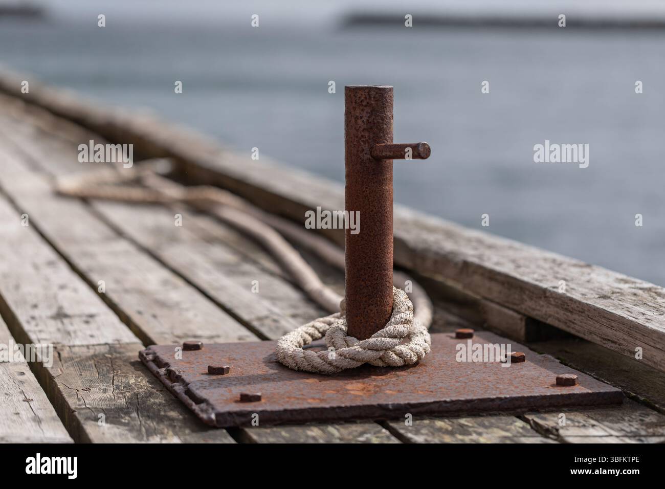 Rostiger Schiffspoller aus Metall mit altem Seil auf verwittertem Holzpier an der Küste der Lofoten in Nordnorwegen. Das Meer und der Pier werden in sanfte Hintergrundunschärfen eingeblendet. Stockfoto