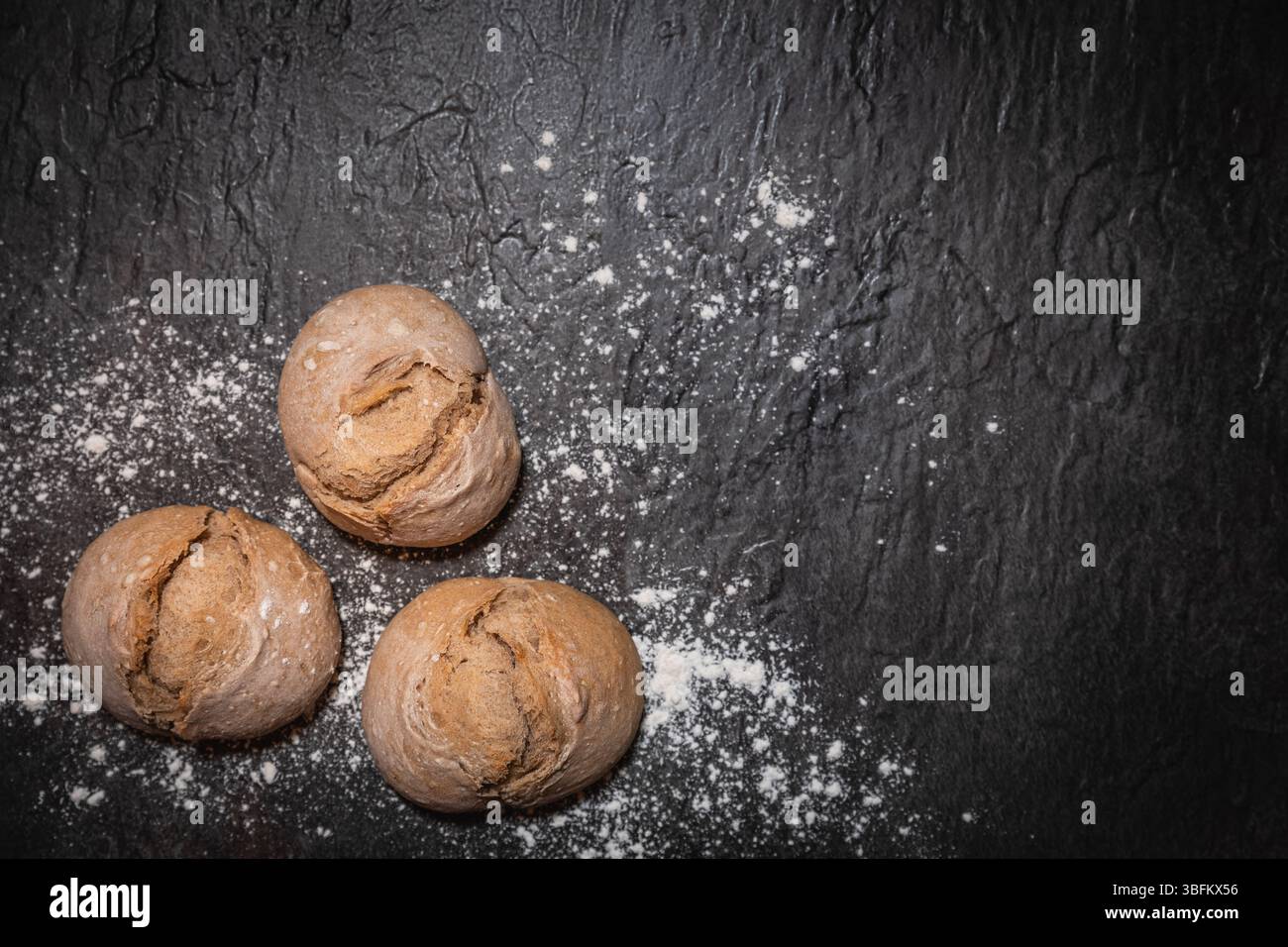 Rustikale Sauerteigbrötchen mit Mehlstaub in der unteren rechten Ecke auf dunklem Hintergrund, Ansicht von oben nach unten. Der Rest des Rahmens lässt Platz für Text Stockfoto