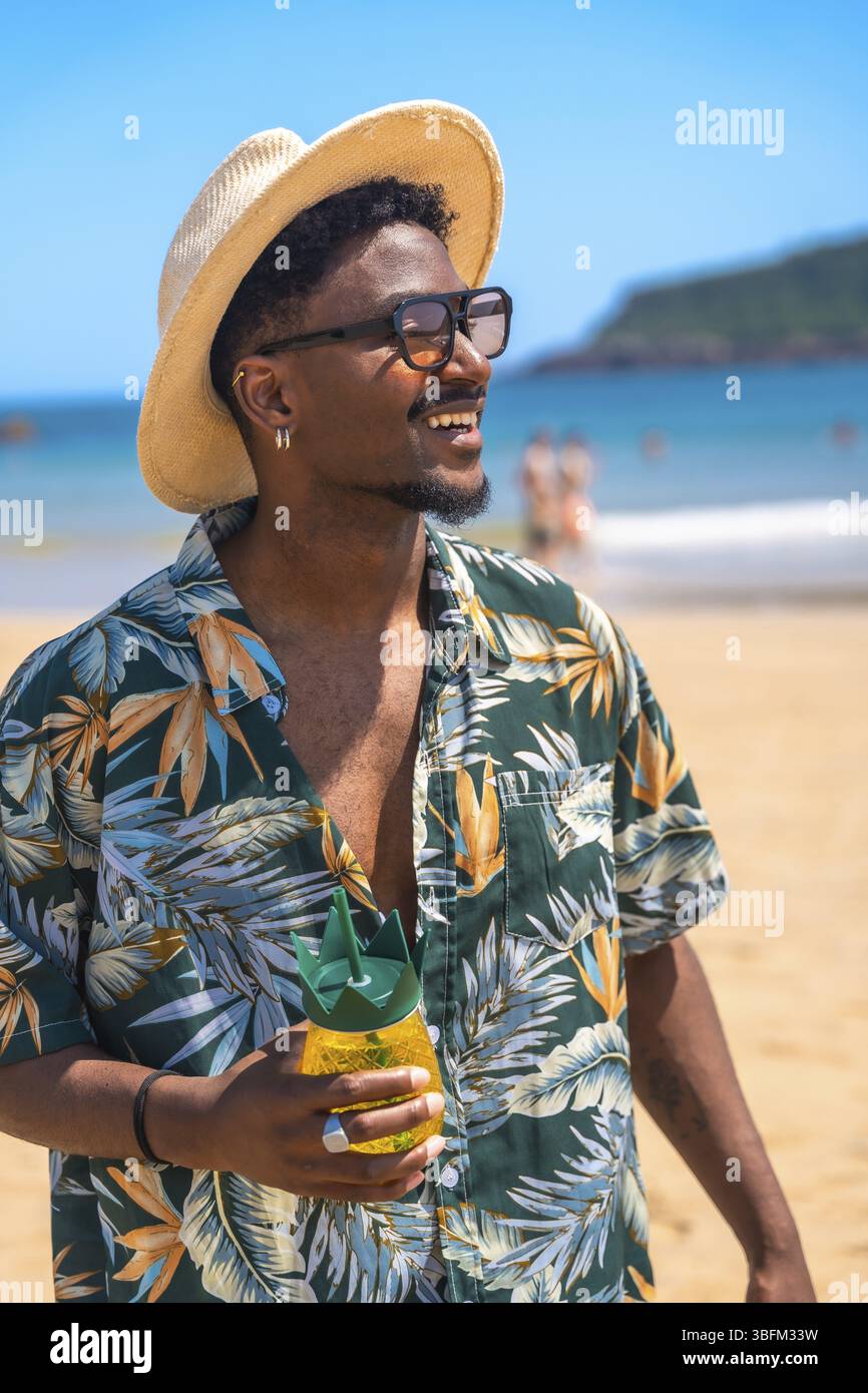 Junger Mann mit Sonnenbrille und Hut, in der Hand eines ananasförmigen Getränks, während er die Sommersonne an einem tropischen Strand genießt Stockfoto