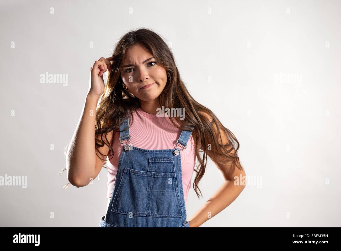 Studioaufnahme in Hüfthöhe, bei der eine junge Erwachsene Frau mit verwirrtem Ausdruck in die Kamera schaut, die eine Hand an der Hüfte und die andere an ihrem Kopf kratzt Stockfoto