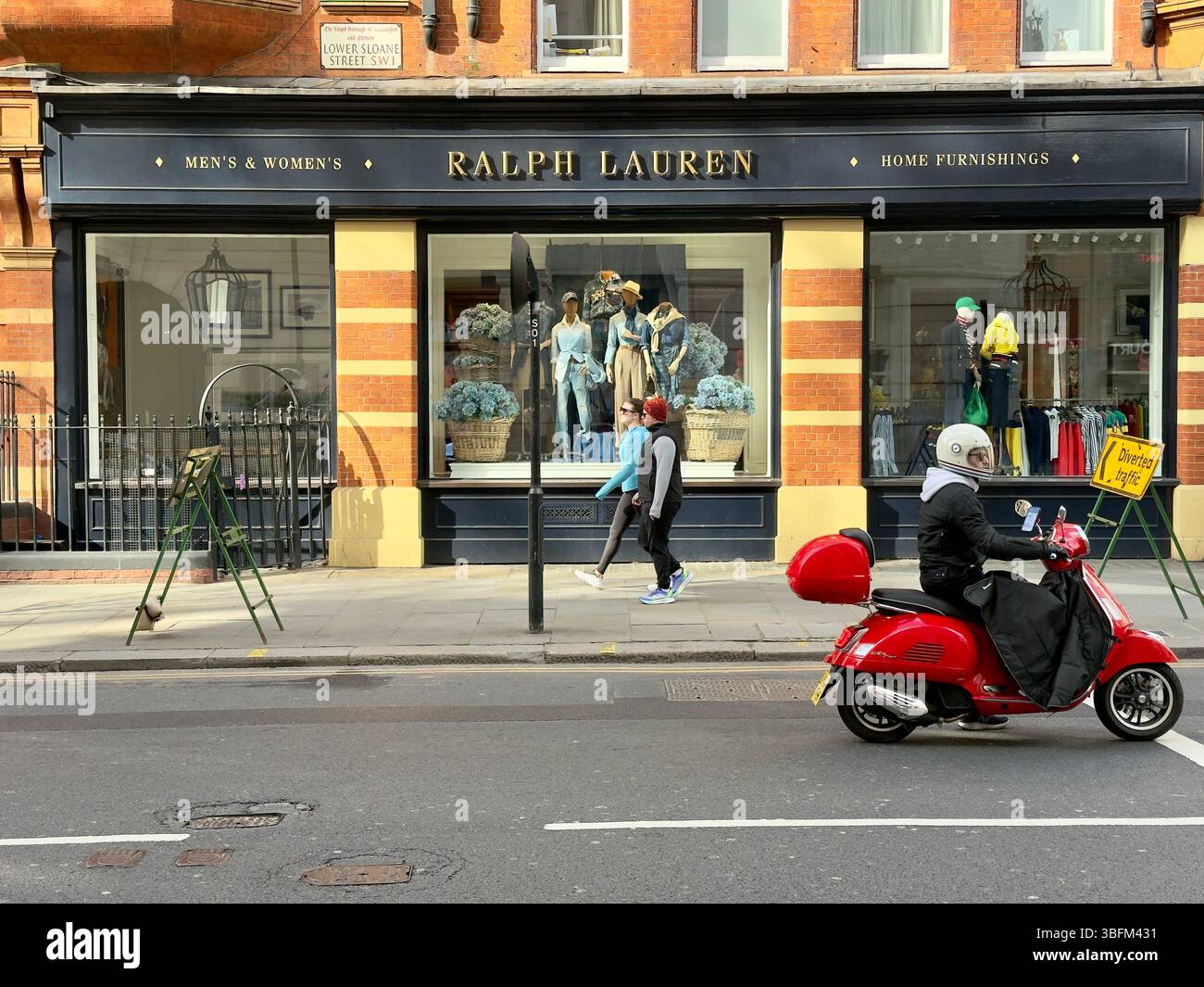LONDON, GROSSBRITANNIEN – 16. MÄRZ 2025: Eine Ralph Lauren-Ladenfront am Sloane Square, London, mit Fußgängern und einem roten Roller, der vorbeifährt. - Smartphone-aufgenommenes Stockfoto