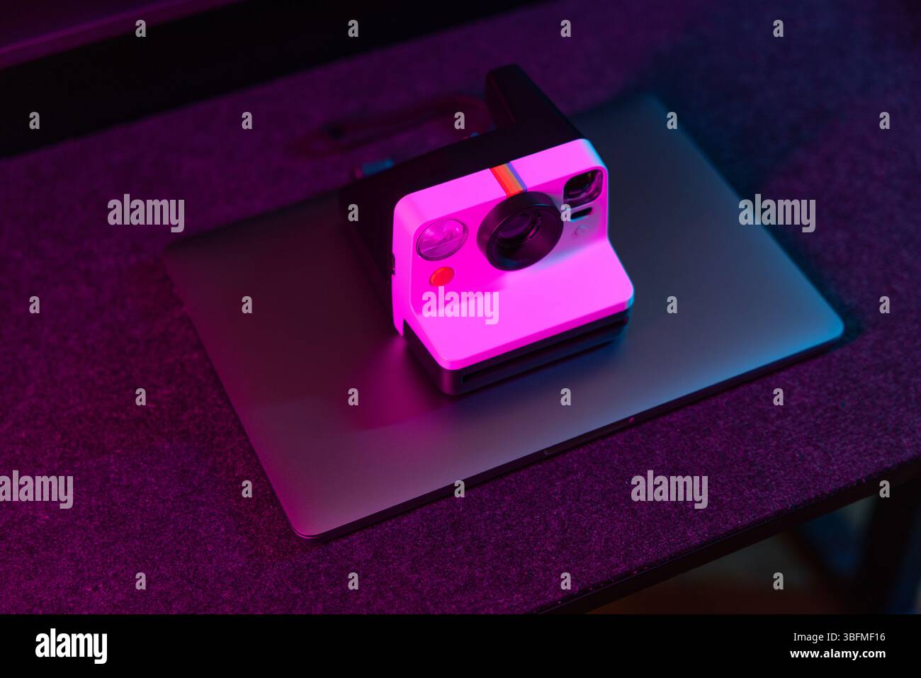 Eine Sofortkamera im Retro-Stil, die auf einem geschlossenen Laptop platziert ist und in leuchtendem Pink und violettem Ambiente auf einem dunklen Schreibtisch liegt. Symbolisiert einen Übergang o Stockfoto