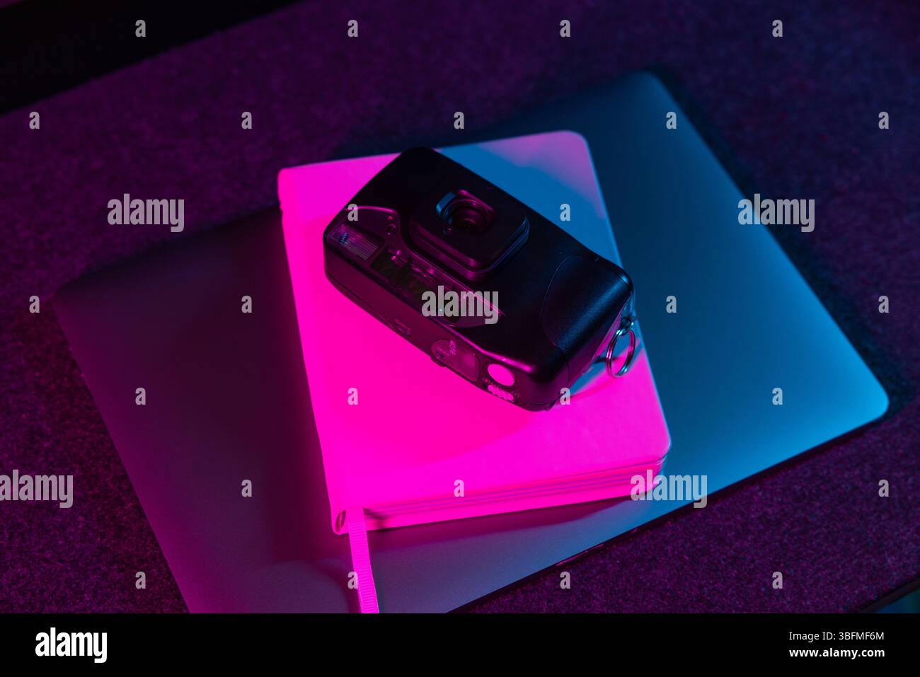 Eine schwarze Point-and-Shoot-Kamera liegt auf einem geschlossenen Notebook auf, das auf einem Laptop gestapelt ist und von leuchtendem rosa und lila Umgebungslicht beleuchtet wird. Stockfoto