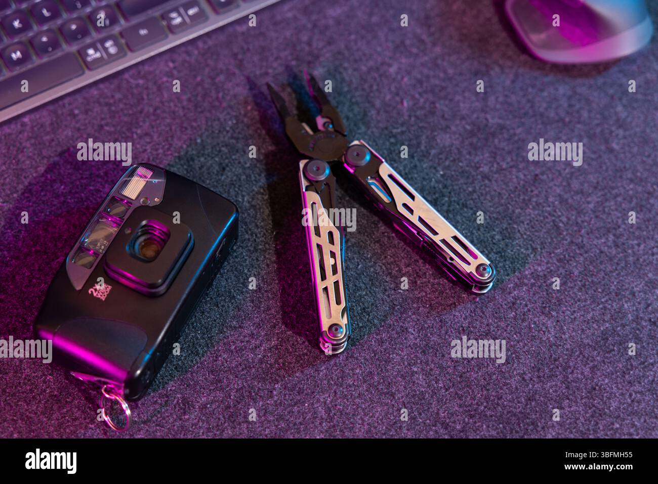 : Flaches Lay-Bild einer Vintage-Filmkamera und ein offenes Multifunktionswerkzeug mit Zange neben einer Computertastatur, unter dramatischem rosa und lila Neonlicht Stockfoto