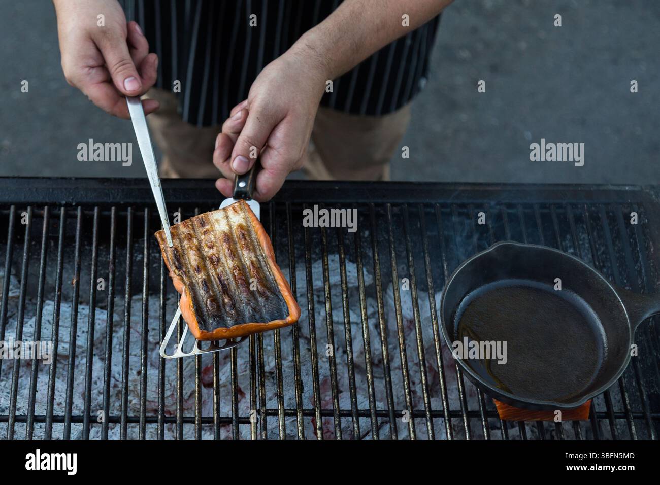 Ein Koch grillt Lachs über einer offenen Flamme, wobei er den Fisch mit einer Zange vorsichtig umdreht, um gleichmäßig zu kochen. Stockfoto