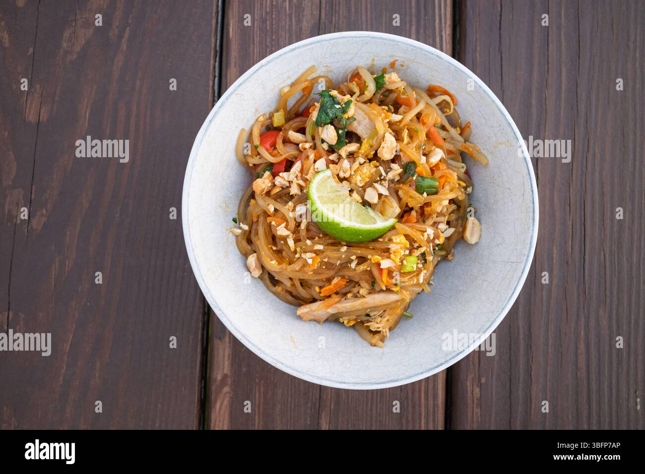 Eine verführerische Schüssel Pad See Ew mit breiten Reisnudeln, Hühnchen, Gemüse und einem frischen Limettenkeil auf Holz. Stockfoto