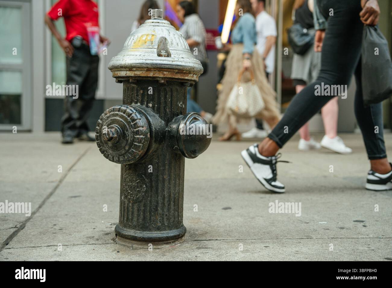 Der Hydrant steht im Fokus, wenn eine Menschenmenge auf dem Bürgersteig der Stadt vorbeiläuft. Urbane Momente in Bewegung. Stockfoto