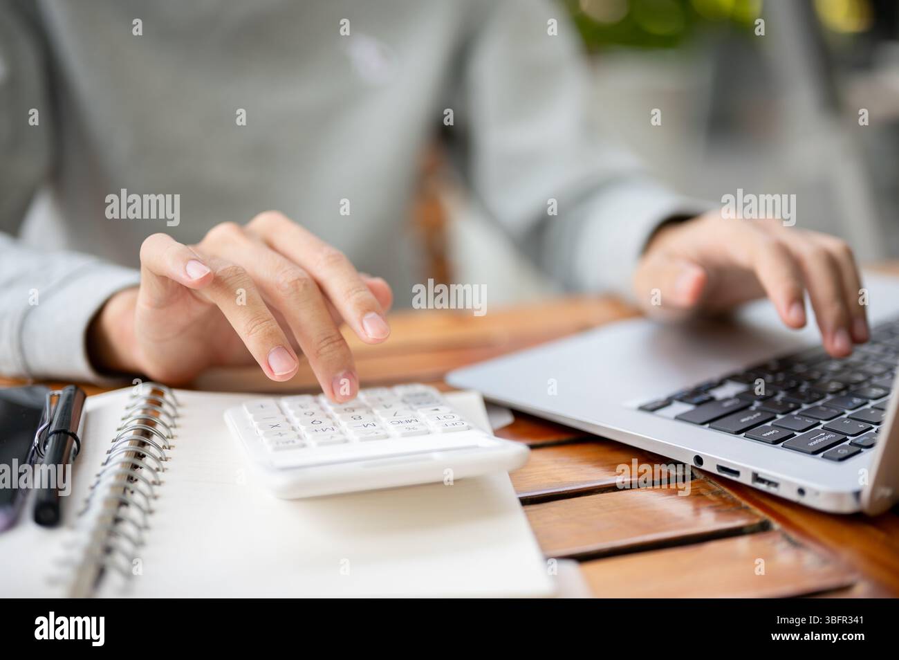 Nahaufnahme einer Hand mit weißem Taschenrechner auf dem Notebook und einer anderen Hand, die auf dem Laptop liegt, während sie am Holztisch sitzt. Im Freien arbeiten oder studieren. Stockfoto