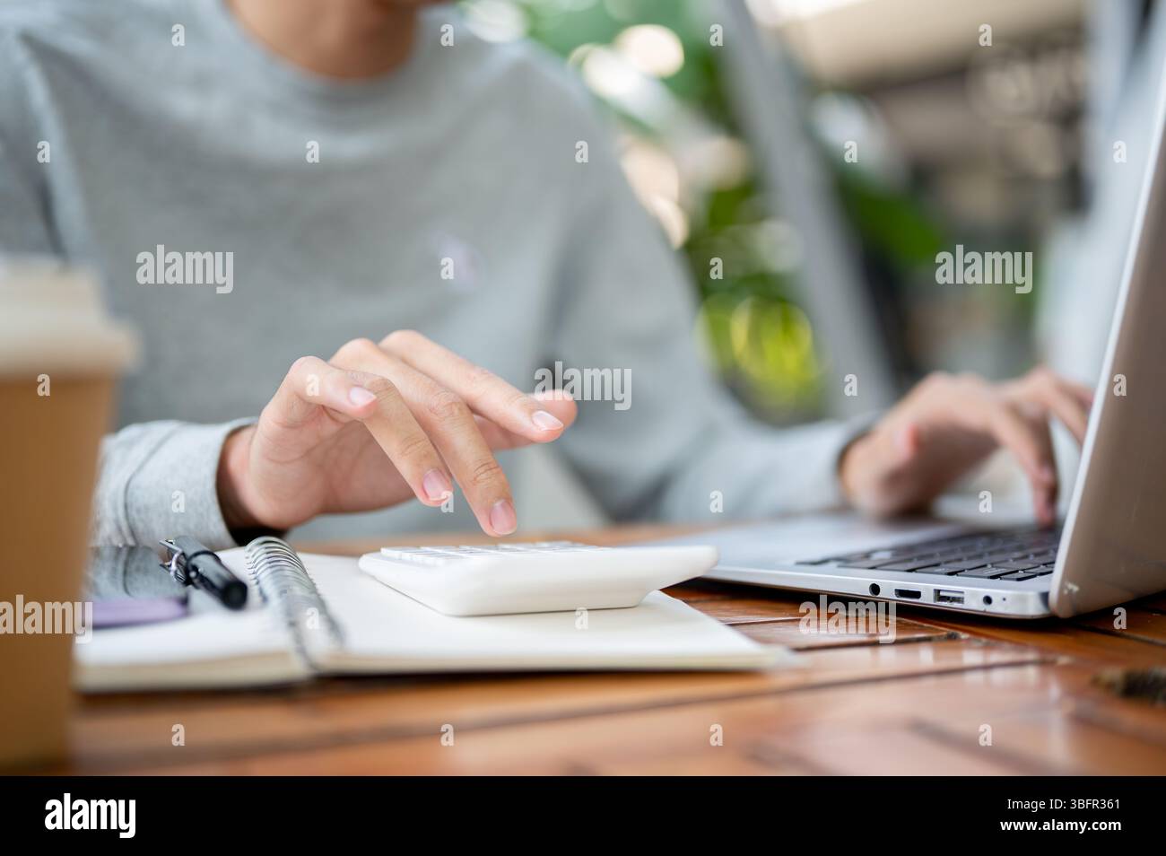 Nahaufnahme einer Hand mit weißem Taschenrechner auf dem Notebook und einer anderen Hand, die auf dem Laptop liegt, während sie am Holztisch sitzt. Im Freien arbeiten oder studieren. Stockfoto