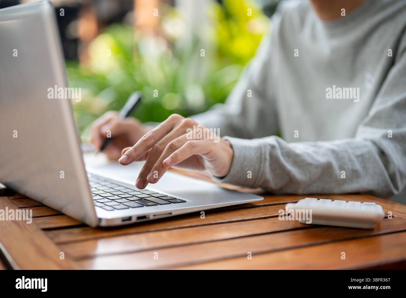Nahaufnahme einer Hand, die einen Stift hält und eine andere Hand auf dem Laptop schreibt, während sie an einem Holztisch in einem Café oder Café sitzt. Im Freien arbeiten oder studieren. Stockfoto