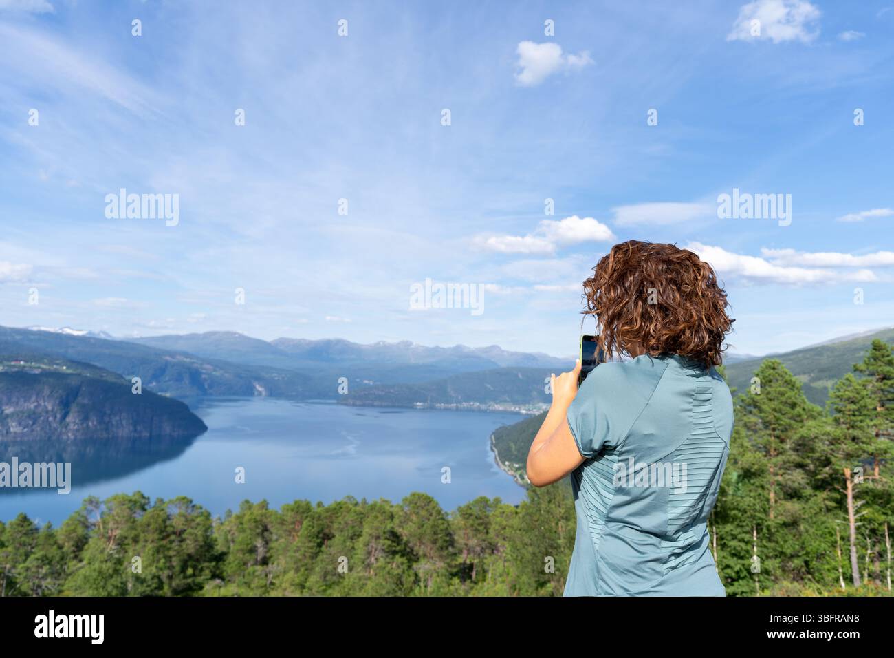 Eine junge Frau in ihren 30ern von hinten gesehen, die an einem sonnigen Sommertag mit ihrem Handy eine atemberaubende norwegische Fjordlandschaft in den Innvikfjorden festnimmt. Stockfoto
