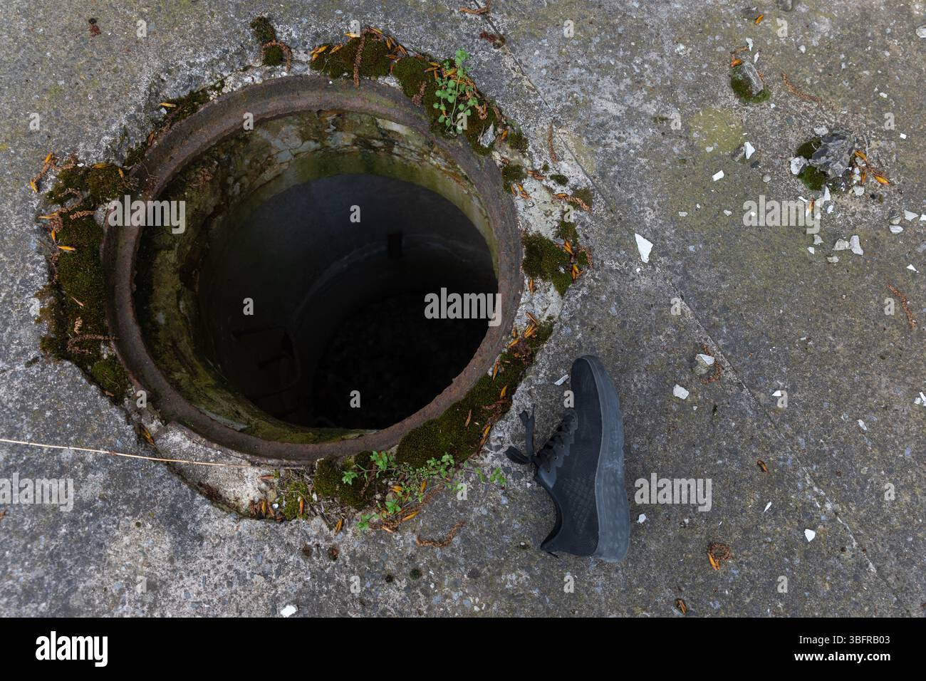 Rostiges rundes Mannloch in gerissenem Beton mit Moos und Trümmern, neben einem einzigen schwarzen Schuh, was auf Verlassenheit oder Rätsel hindeutet. Stockfoto Rostiges rundes Mannloch in gerissenem Beton mit Moos und Trümmern, neben einem einzigen schwarzen Schuh, was auf Verlassenheit oder Rätsel hindeutet. Stockfoto