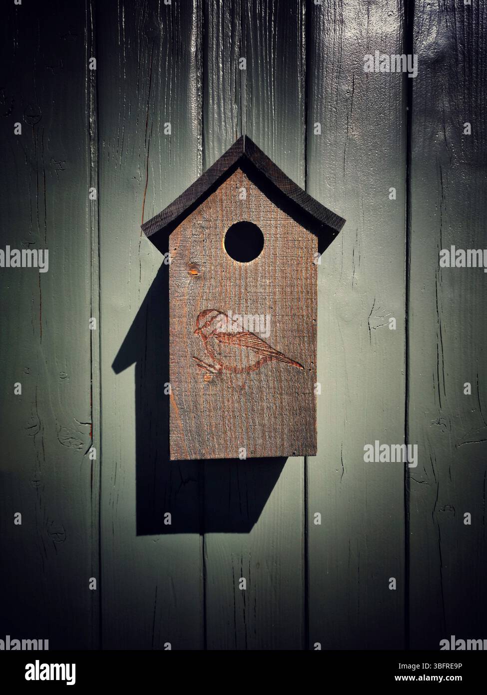 Ein hölzernes Vogelhaus mit geschnitztem Vogelmotiv hängt an einer rustikalen grauen Wand. - Smartphone-aufgenommenes Stockfoto