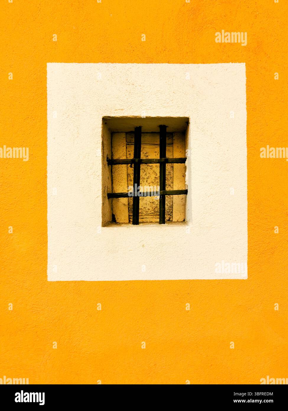 Ein kleines rechteckiges Fenster mit schwarzem Gitter ist in einem weißen Quadrat zentriert, umgeben von einer leuchtend gelben Wand. Stockfoto
