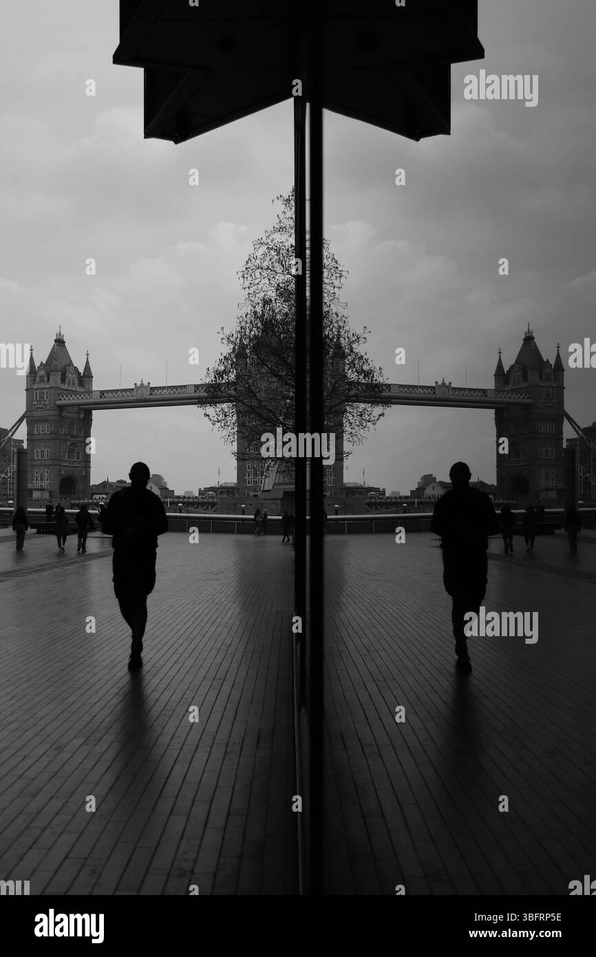 Eine Einzelfigur geht an einer reflektierenden Glaswand in London vorbei und schafft einen Spiegelblick auf die Tower Bridge unter einem stimmungsvollen Himmel in Schwarz-weiß-Tönen Stockfoto