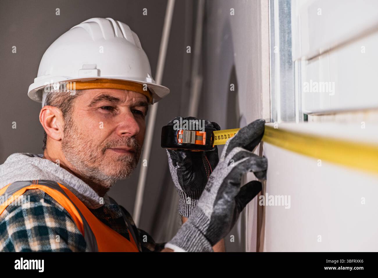 Fokussierter Bauarbeiter, der ein Maßband verwendet, um genaue Abmessungen bei der Wandmontage auf der Baustelle sicherzustellen Stockfoto