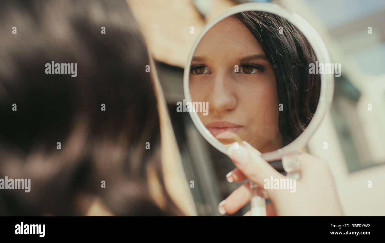 Eine junge Frau schaut in einen Handspiegel, während sie Make-up aufträgt. Sie ist modisch und posiert vorsichtig für einen bevorstehenden Social-Media-Beitrag. Stockfoto