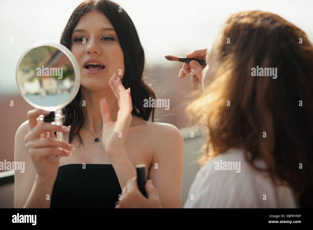 Eine junge Frau in einem schwarzen Kleid hält einen Spiegel und überprüft ihr Gesicht. Ein Make-up-Künstler trägt Lippenstift mit einem Pinsel auf. Es scheint, als wären sie auf einem Dach Stockfoto