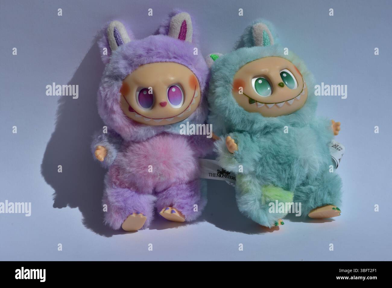 Zwei verspielte Labubu-Spielzeugfiguren in flauschigen blau-rosa Monsterkostümen und einem schelmischen Lächeln vor weißem Hintergrund. Stockfoto
