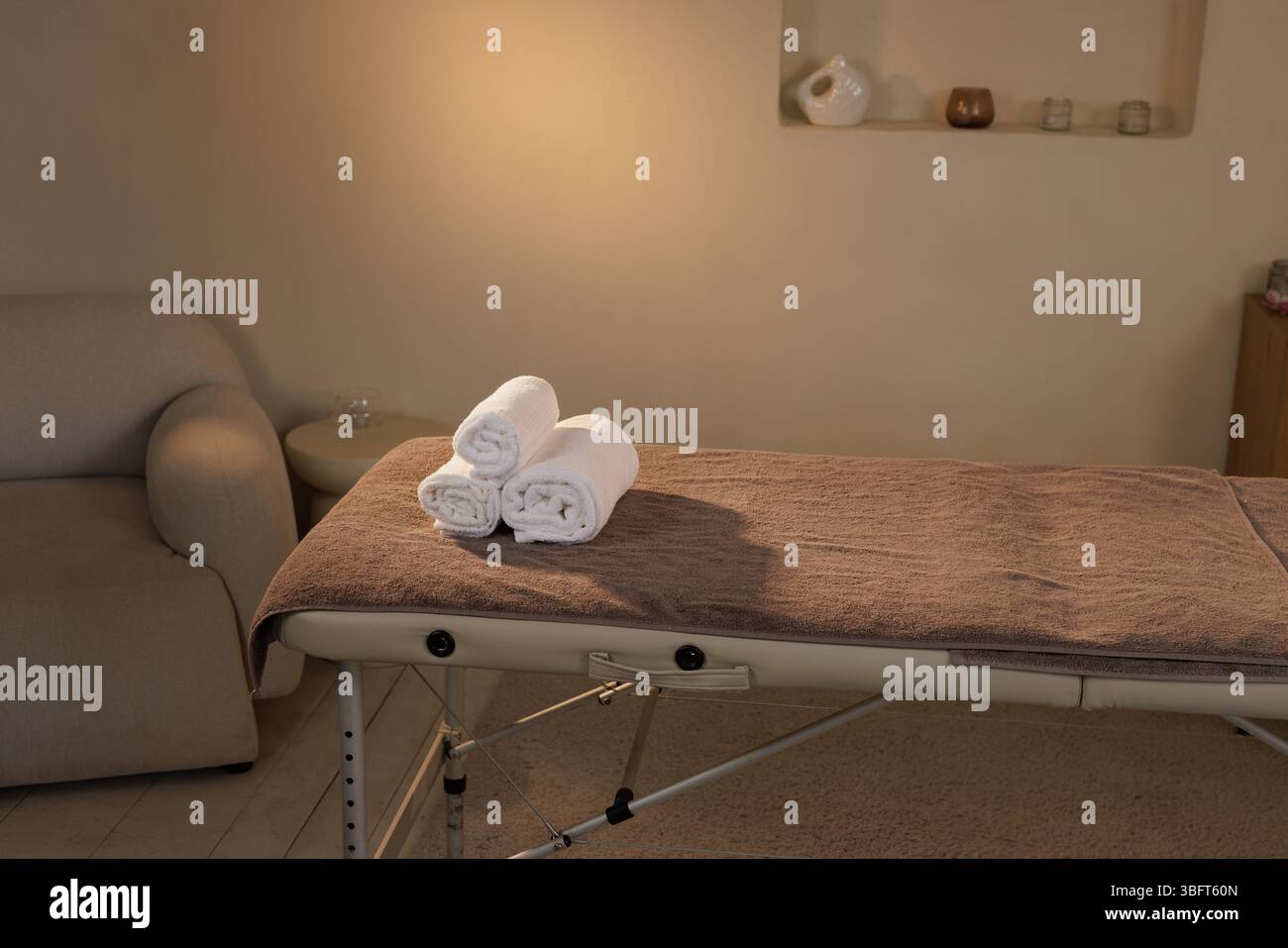Massagetisch mit braunem Handtuchbezug und weißen gerollten Handtüchern in ruhiger Spa-Einrichtung, bereit für Therapie oder Behandlung. Stockfoto
