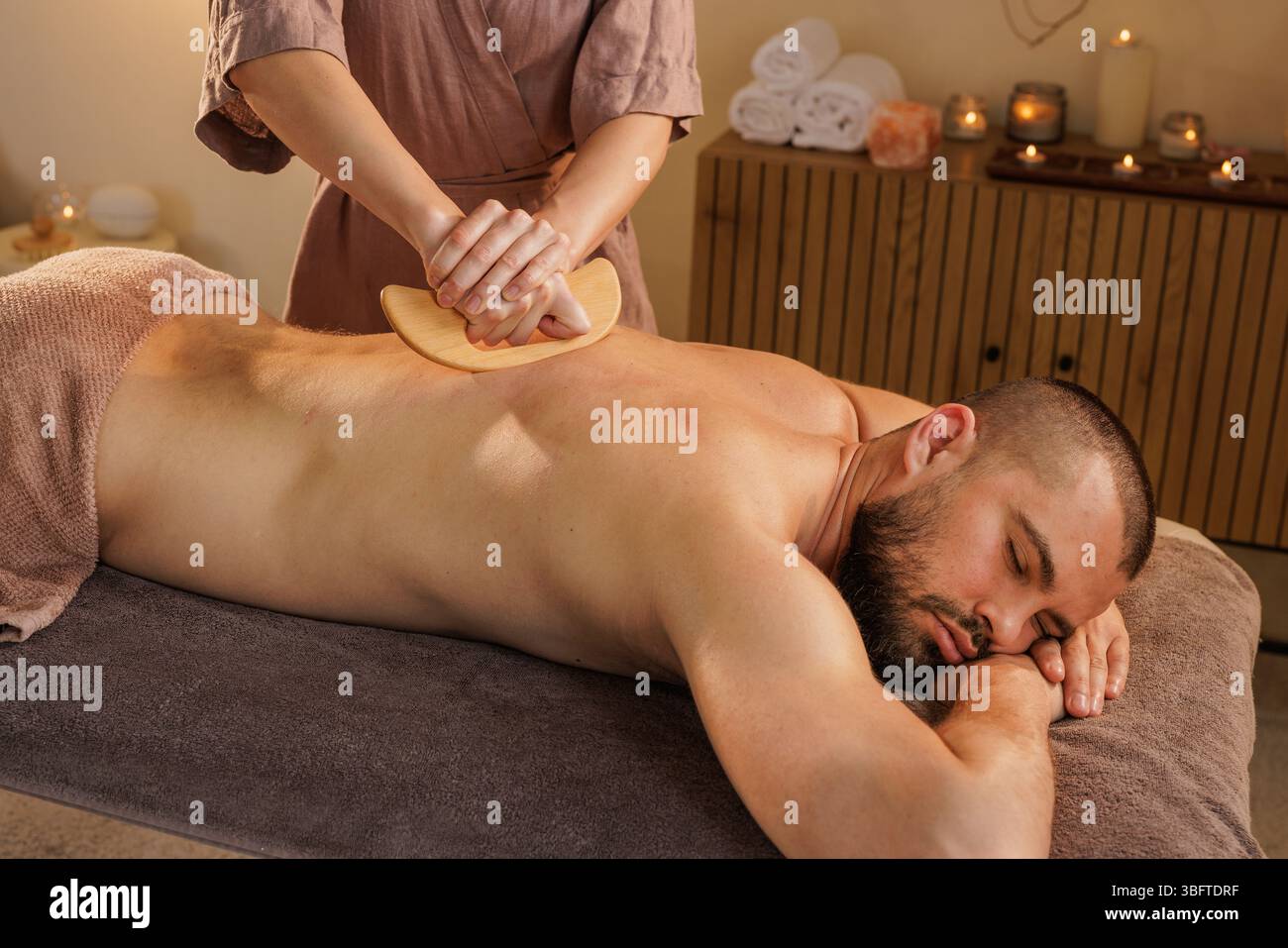 Der Therapeut verwendet ein hölzernes Massagewerkzeug für eine Tiefenmassage des Rückengewebes bei einem entspannten Mann in einer ruhigen Spa-Umgebung mit Kerzen und Handtüchern. Stockfoto