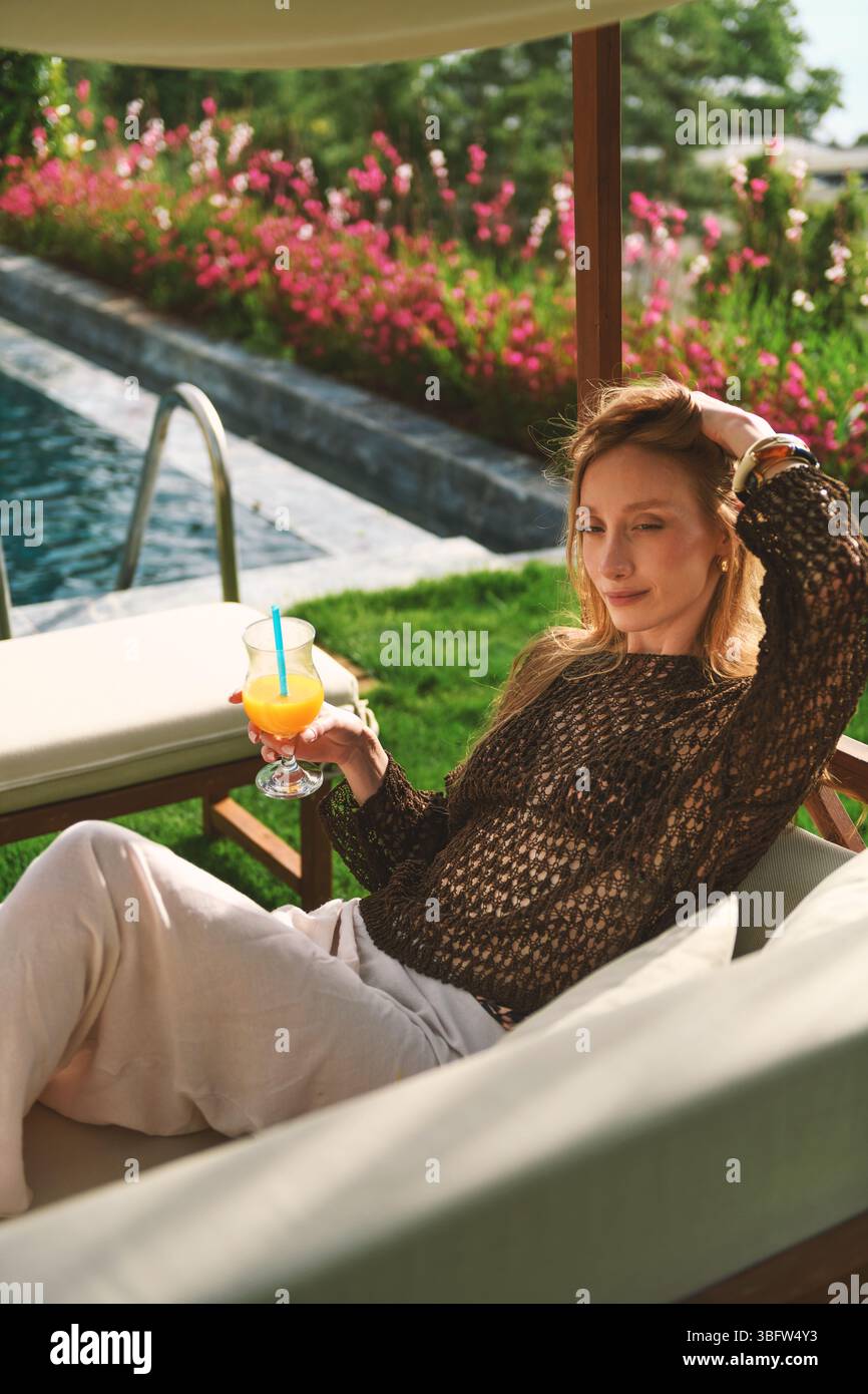 Eine blonde Frau aus den 20er Jahren genießt Orangensaft, während sie im privaten Garten ihres Hotels posiert, wobei der Pool hinter ihr sichtbar ist. Stockfoto
