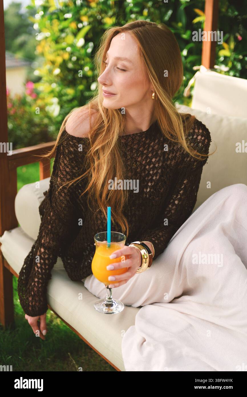 Eine blonde Frau aus den 20er Jahren sitzt auf einem Gartenstuhl, schlürft Orangensaft und lächelt im privaten Garten ihres Hotels für die Kamera. Stockfoto