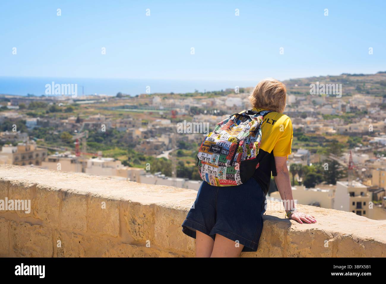 Alleinreisender, Rückansicht einer jungen Reisenden, die sich an eine alte Steinbrüstung lehnt und auf eine mediterrane Stadt, Gozo, Malta, blickt Stockfoto