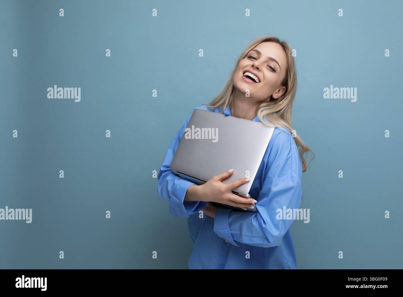 Lächelnde, fröhliche blonde Schülerin mit einem Laptop auf einem hellblauen Hintergrund Stockfoto