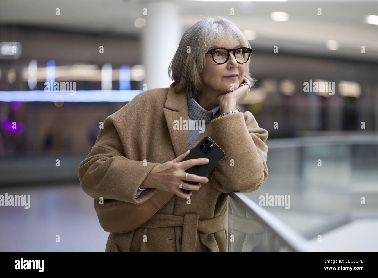 Eine Frau mit stilvoller Brille lehnt sich an ein Geländer und hält nachdenklich ihr Smartphone. Sie scheint sich in einer zeitgenössischen Umgebung verloren zu haben Stockfoto