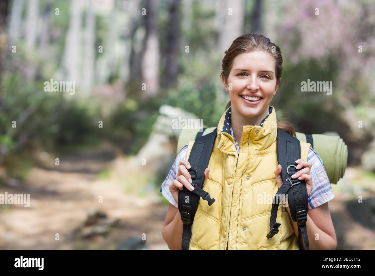 Frau, die auf dem Waldweg steht, gelbe Weste trägt und Wanderrucksack mit Schlafmatte hält Stockfoto