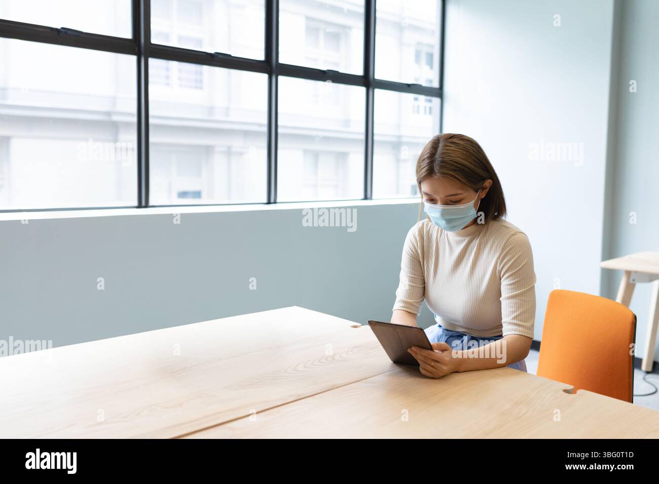 Asiatische Frau mit Gesichtsmaske und blassem Oberteil, die am Schreibtisch sitzt und Tablette in der Nähe des Gitterfensters hält Stockfoto
