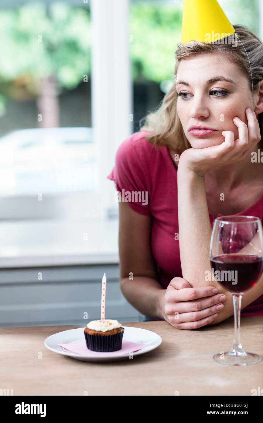 Frau mit gelbem Partyhut, die am Fenstertisch sitzt, Cupcake und Wein ansieht, Kopierraum Stockfoto