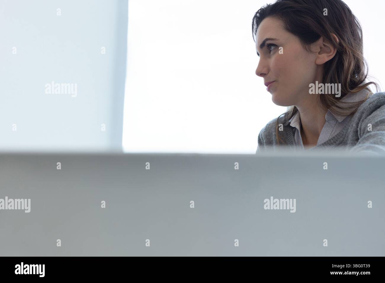 Frau, die hinter dem grauen Schreibtisch sitzt, Hemd mit Kragen und Pullover im Büro mit Fenster und Kopierraum trägt Stockfoto
