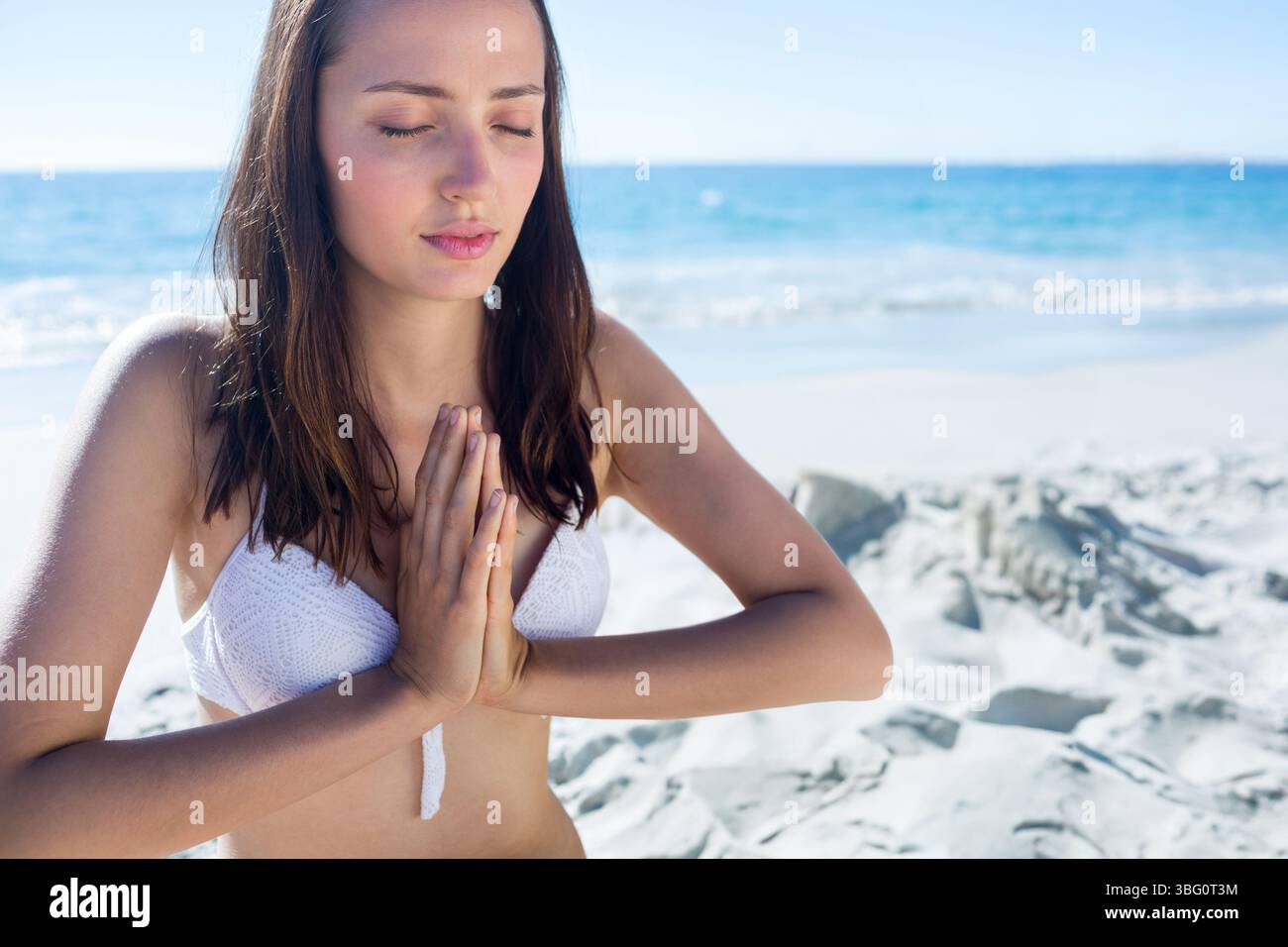 Meditierende Frau, die am Sandstrand am Strand sitzt, in weißem Bikinioberteil unter hellem Sonnenlicht Stockfoto