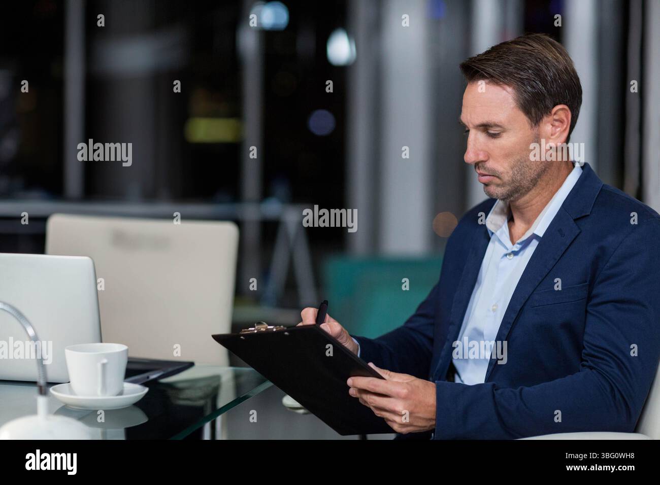 Geschäftsmann sitzt am Schreibtisch mit Glasplatte im Büro und macht Notizen auf der Zwischenablage in der Nähe einer Laptop-Kaffeetasse Stockfoto