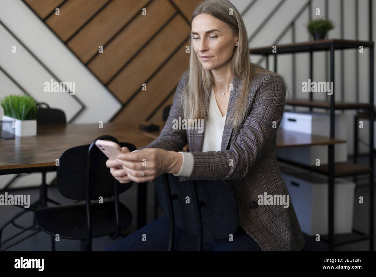 Eine Frau im Blazer sitzt an einem Holztisch in einem modernen Büro. Sie konzentriert sich auf ihr Smartphone, umgeben von minimalistischem Dekor und Pflanzen im Innenbereich Stockfoto