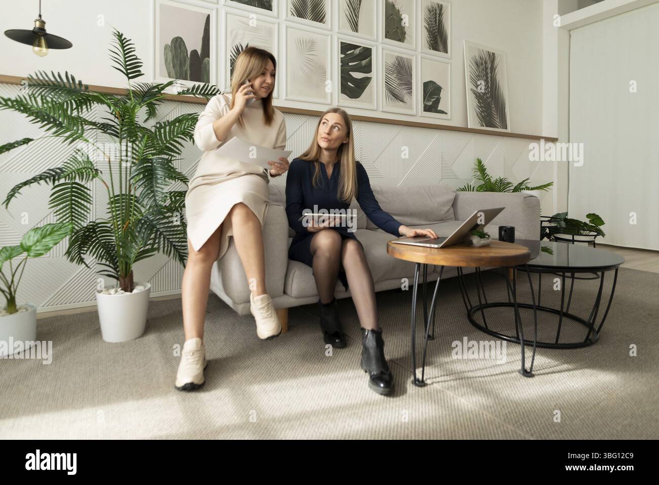Zwei Frauen nehmen an einer Arbeitsdiskussion in einem modernen Bürobereich Teil, umgeben von Pflanzen und Kunstwerken. Eine Frau gibt Gesten, während die andere bei beobachtet Stockfoto