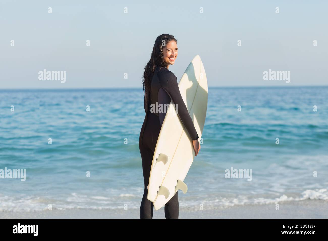 Weibliche Surferin in schwarzem Neoprenanzug lächelt, während sie ein weißes Surfbrett am Sandstrand hält Stockfoto