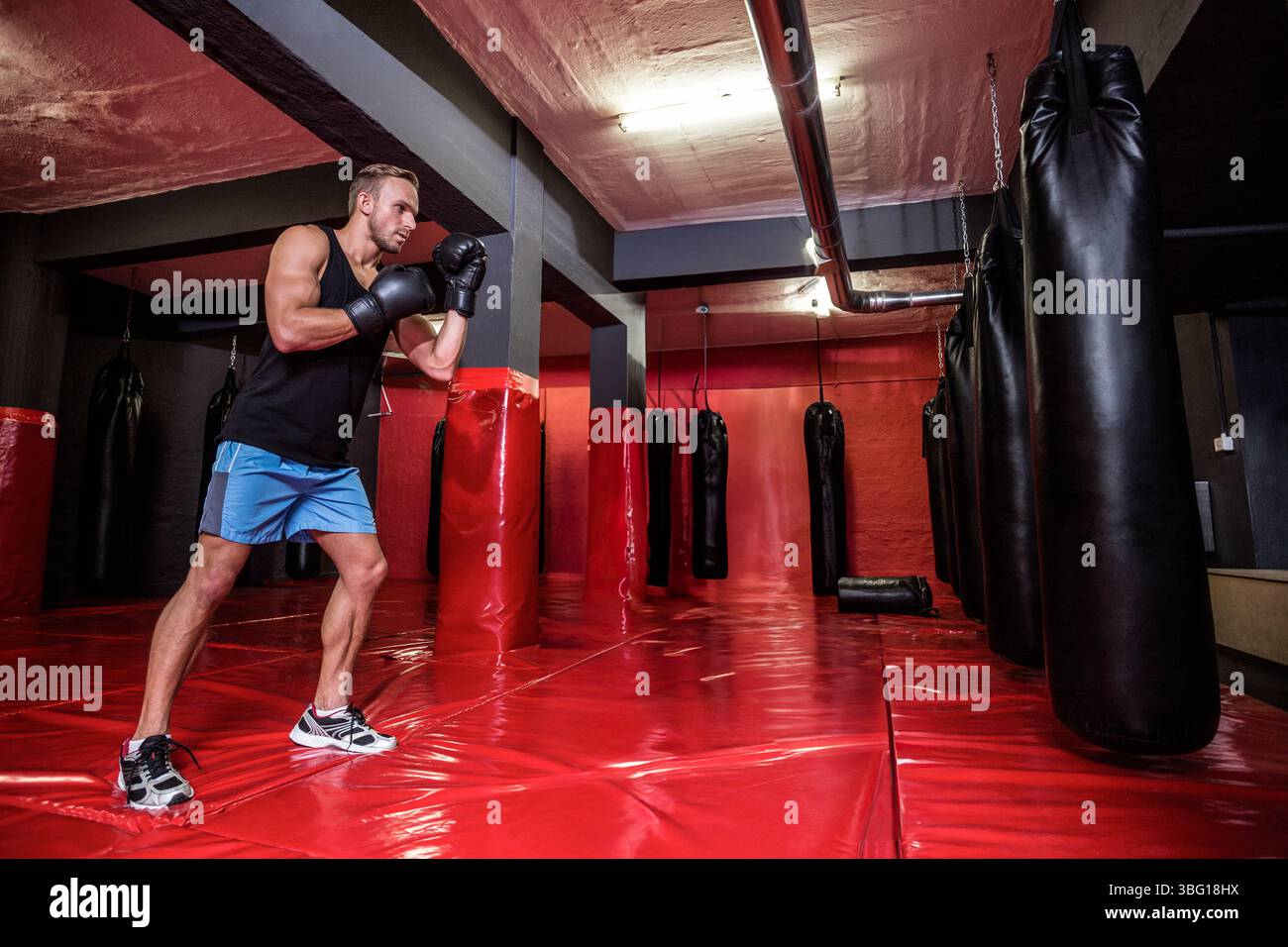 Mann, der Boxen im Keller des Fitnessstudios übt und schwere Tasche mit Handschuhen auf roten Matten schlägt, Kopierraum Stockfoto