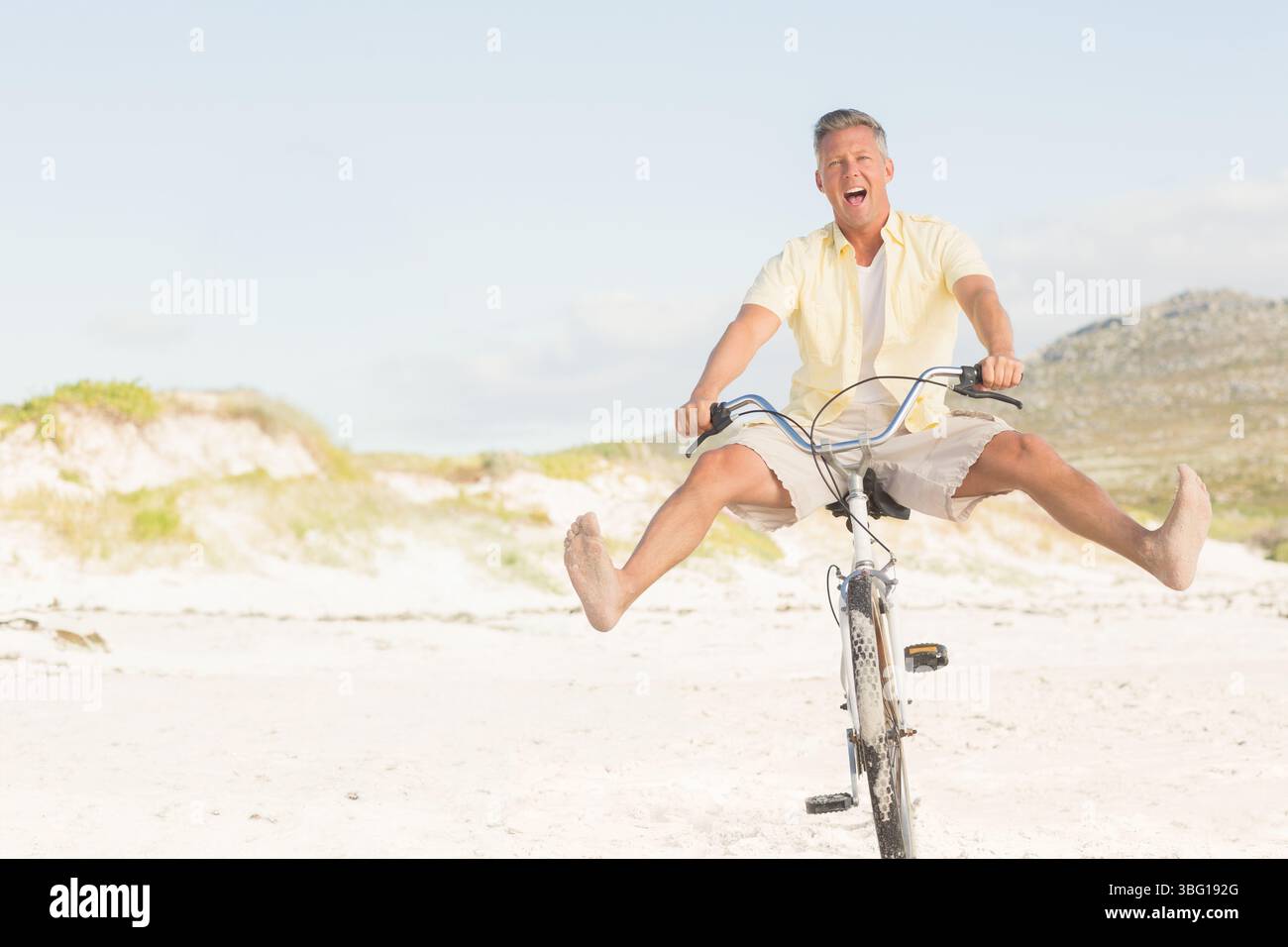 Mann mittleren Alters, der mit dem Strandkreuzfahrrad entlang der Sanddünen fährt, während er die Beine hochtritt und lacht Stockfoto