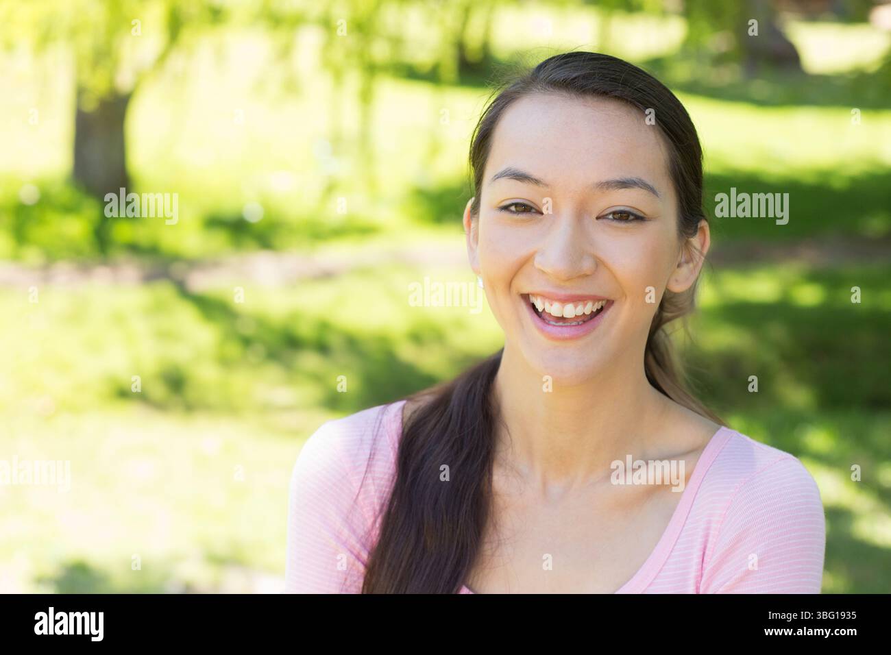 Asiatische Frau, die im Park steht, mit rosafarbenem Oberteil zwischen grünem Gras und Bäumen, Kopierraum Stockfoto