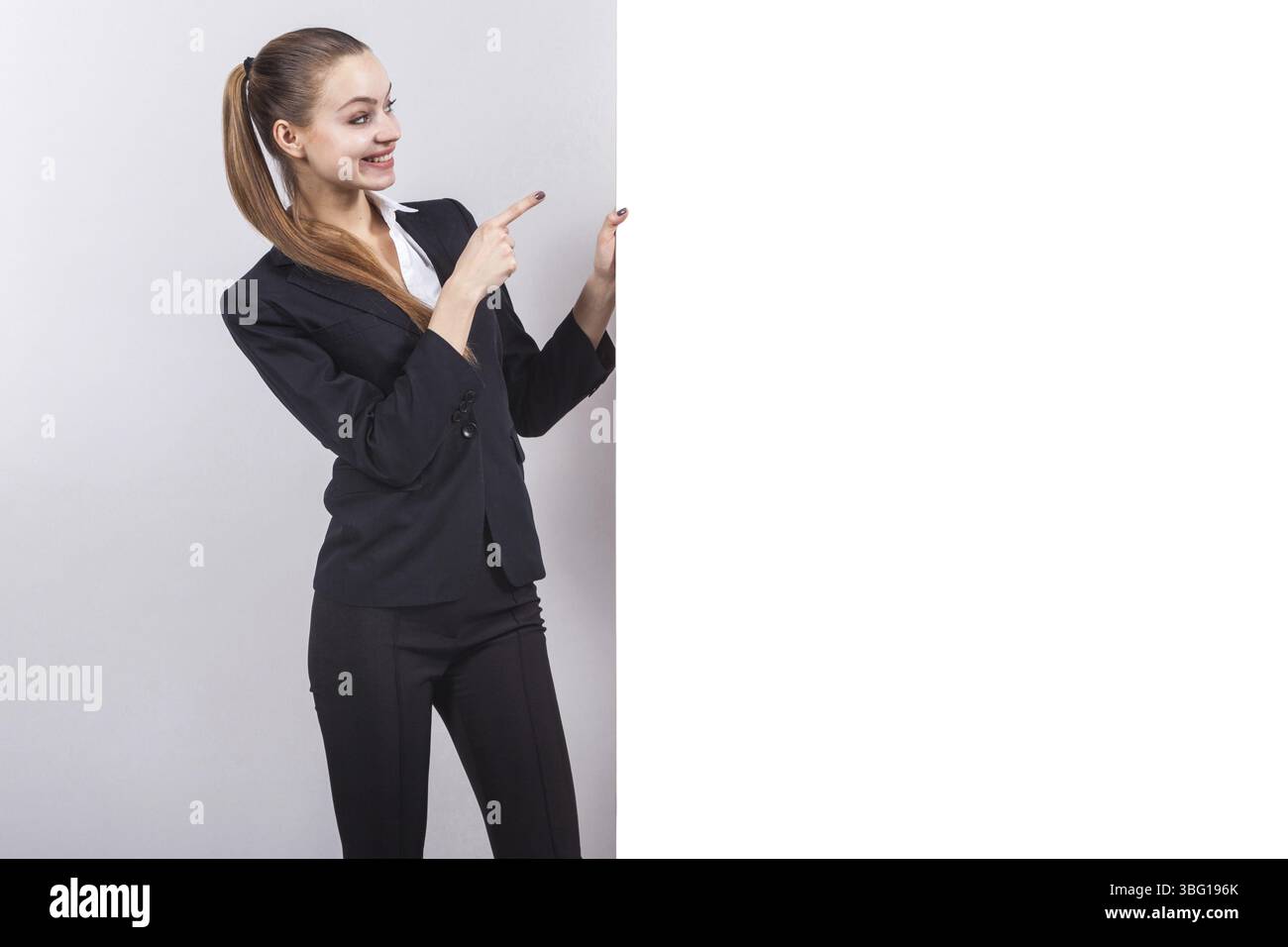 Junge attraktive kaukasische Geschäftsfrau mit Pferdeschwanz in schwarzem Anzug, Blick auf leere Seite isoliert auf grauem, leerem Teil des Bildes Stockfoto