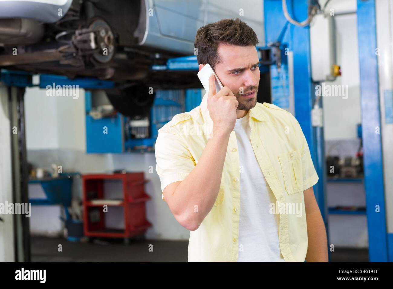 Mann in Zwanzigern, der das Smartphone hält, während er in der Reparaturhalle mit erhöhtem Aufzug steht, Kopierraum Stockfoto