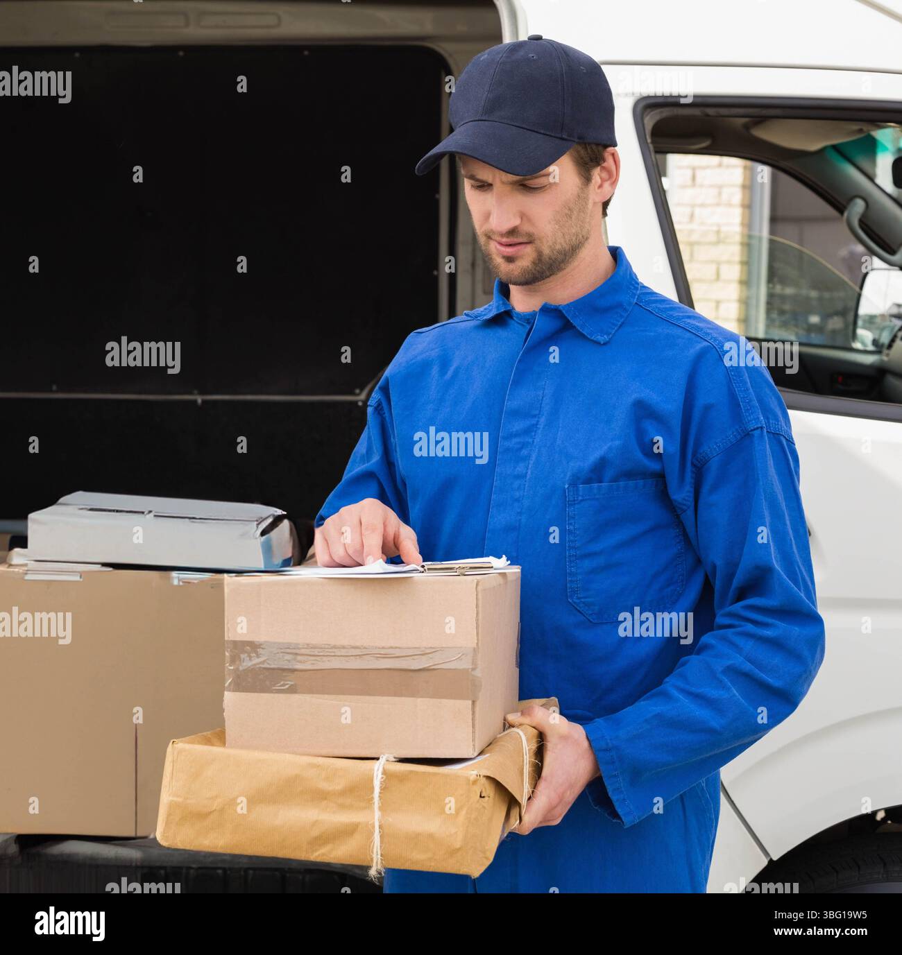 Männlicher Kurier in Uniform, der die Zwischenablage überprüft und Pakete neben dem weißen Van auf einer gepflasterten Einfahrt hält Stockfoto