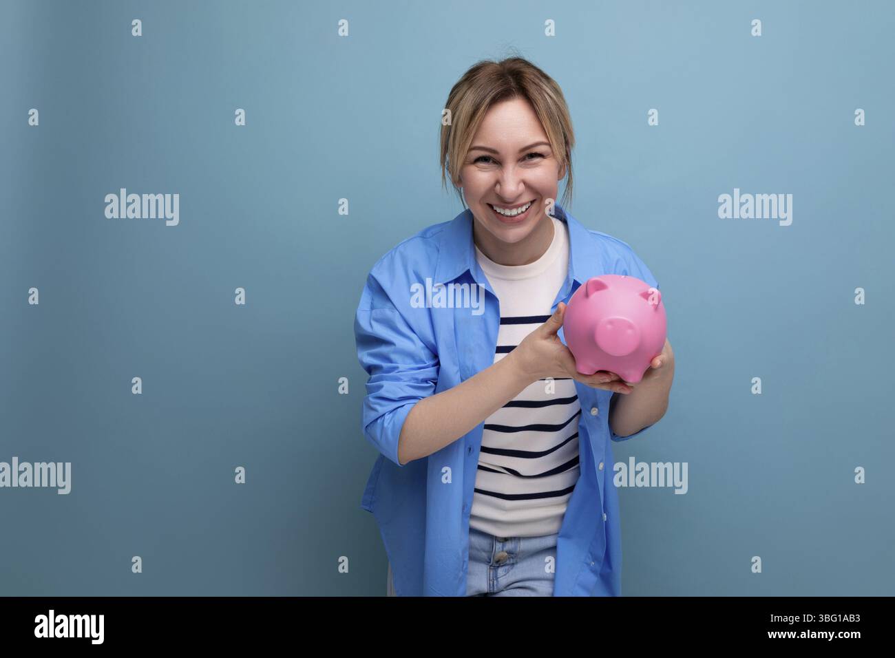 Blonde attraktive junge Frau in lässiger Kleidung mit einem Lächeln, die eine Sparkasse mit Ersparnissen in den Händen auf einem blauen Hintergrund mit Kopierraum hält Stockfoto