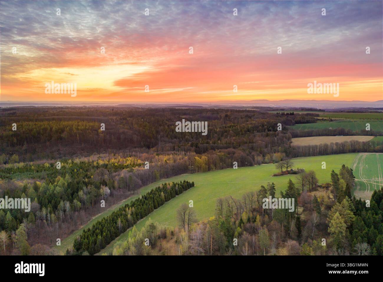 Von oben aus der Drohne blickt man auf einen Herbstwald bei Sonnenuntergang, Nadel- und Laubbäume in Grün-, Orange- und Brauentönen mit orangen Wolken Stockfoto