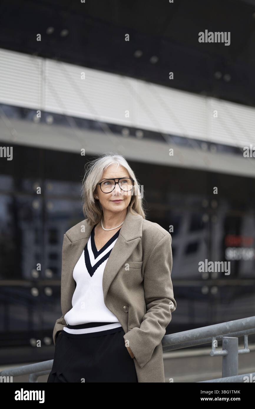 Eine ältere Geschäftsfrau steht vor ihrem Büro, in einem schicken Outfit gekleidet, und sieht selbstbewusst und entspannt in der Mittagssonne aus, während sie nachdenklich schaut Stockfoto